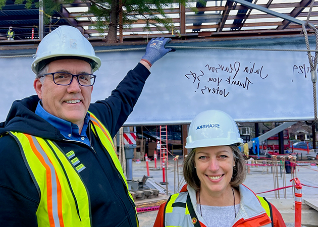 a short person and a tall person wear construction safety vests and hard hats while standing in front of part of a steel beam painted white. handwriting on the beam says "thank you SPS voters, Julia Pearson"