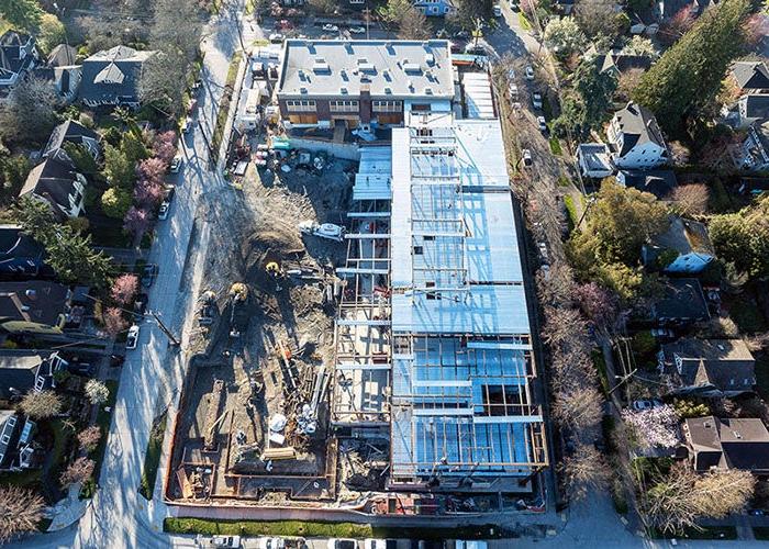 aerial view of a construction site with a brick building at the top and a new building being constructed at a right angle on the left side. a foundation is in progress in the lower right corner of the site dirt and construction equipment are in the remaining space